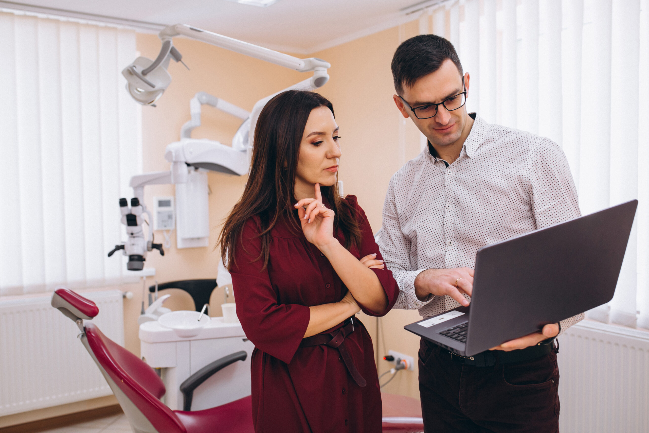 A man and woman in a dental office discuss information on a laptop. The woman appears thoughtful, while the man gestures at the screen. Dental chair nearby.