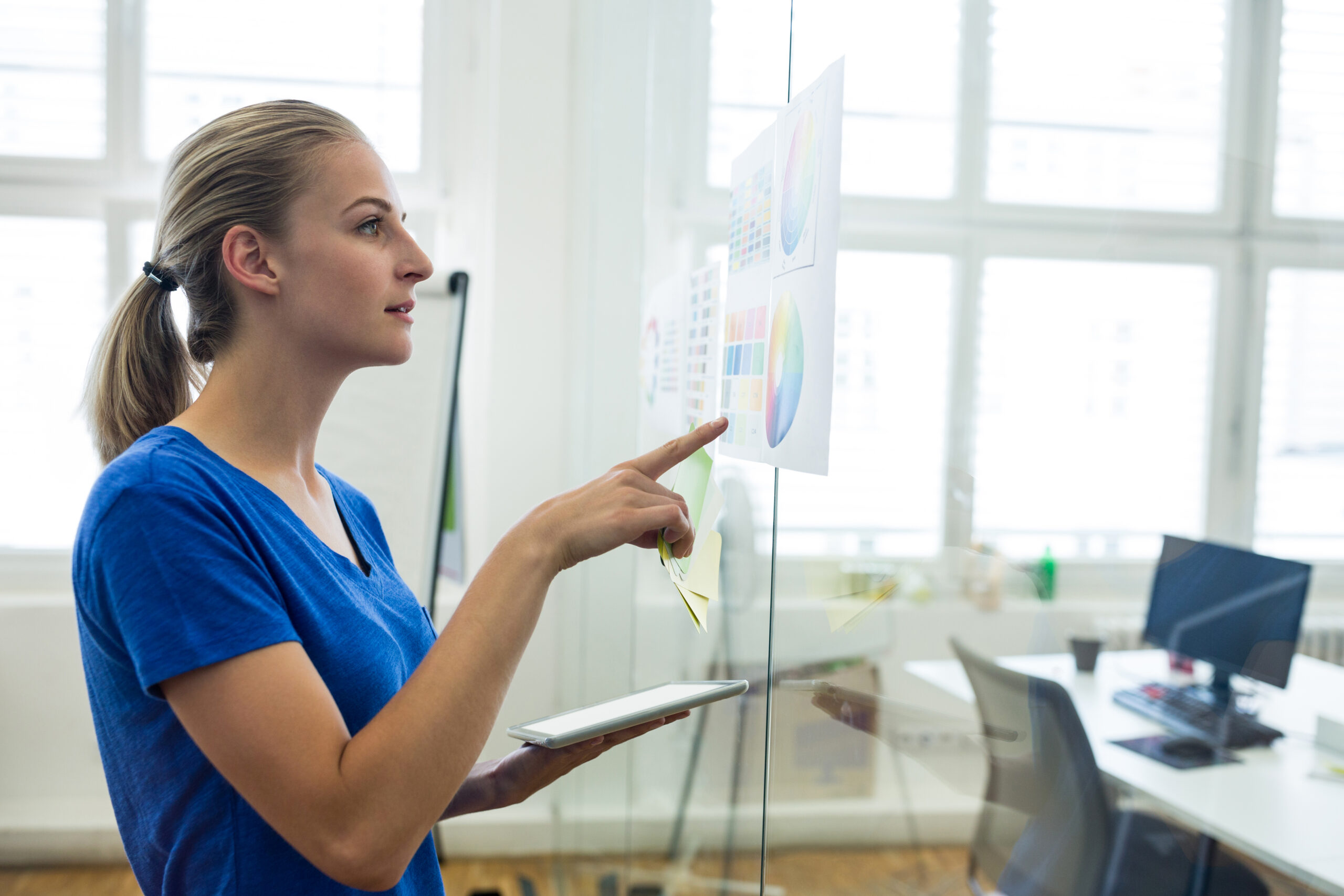 Woman in blue shirt holding a tablet, pointing at charts on glass wall. Bright office setting, work-focused and analytical atmosphere.