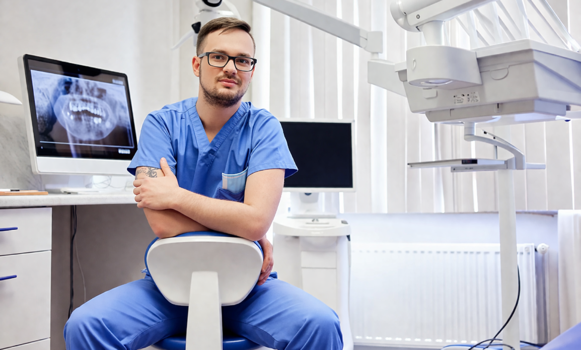 En tandläkare i blå arbetskläder sitter självsäkert på en stol i en modern tandklinik. En röntgenbild syns på en bildskärm, och tandvårdsutrustning omger honom.