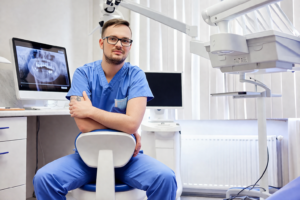 En tandläkare i blå arbetskläder sitter självsäkert på en stol i en modern tandklinik. En röntgenbild syns på en bildskärm, och tandvårdsutrustning omger honom.