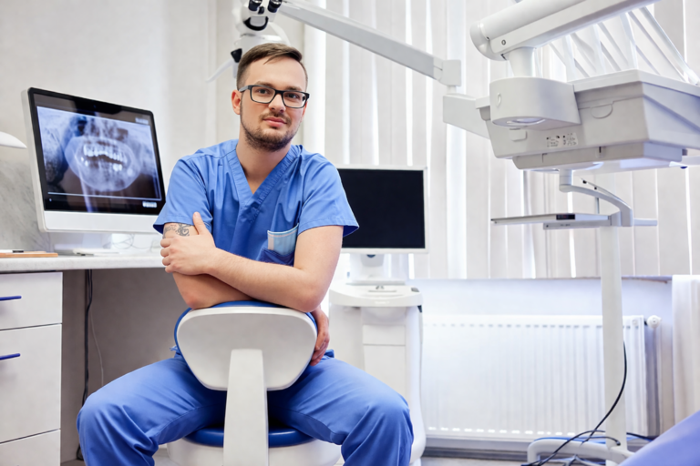 En tandläkare i blå arbetskläder sitter självsäkert på en stol i en modern tandklinik. En röntgenbild syns på en bildskärm, och tandvårdsutrustning omger honom.