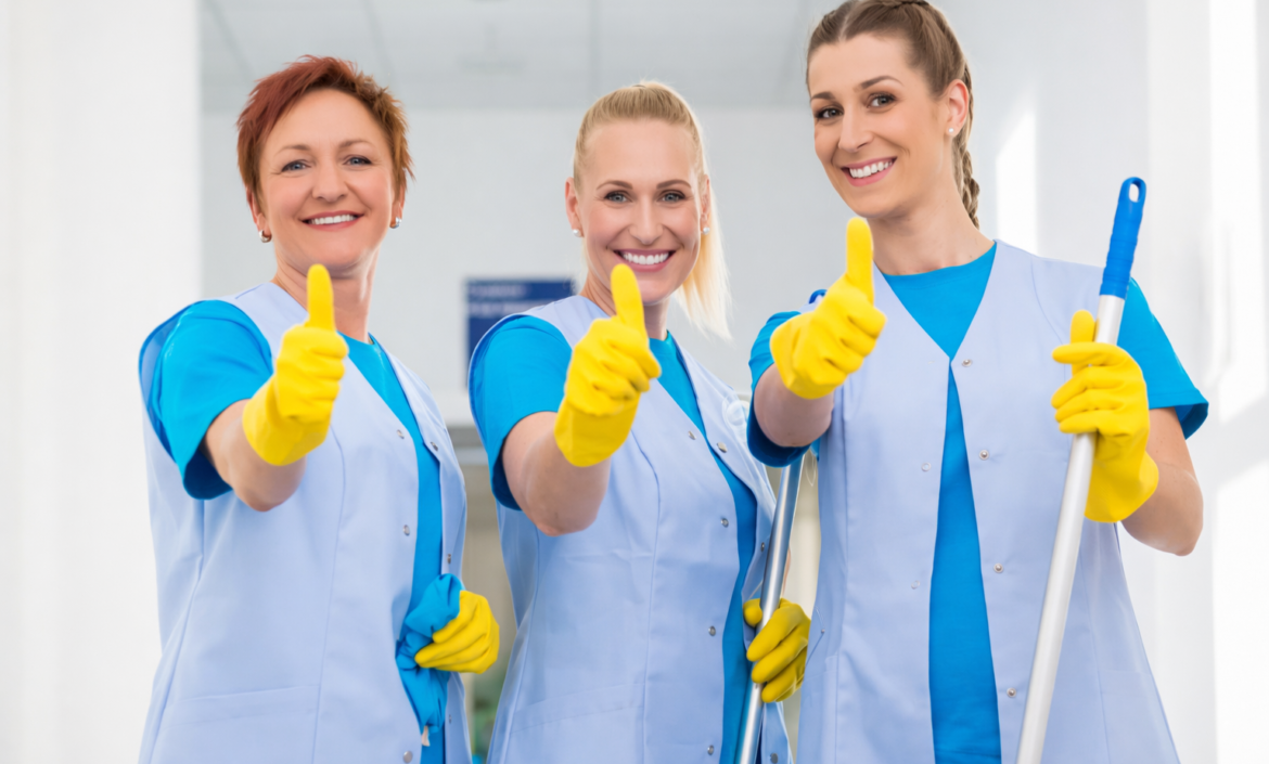 Tre kvinnor i blå uniformer och gula handskar ler och ger tummen upp. De står i en ljus hall och utstrålar positivitet och lagarbete