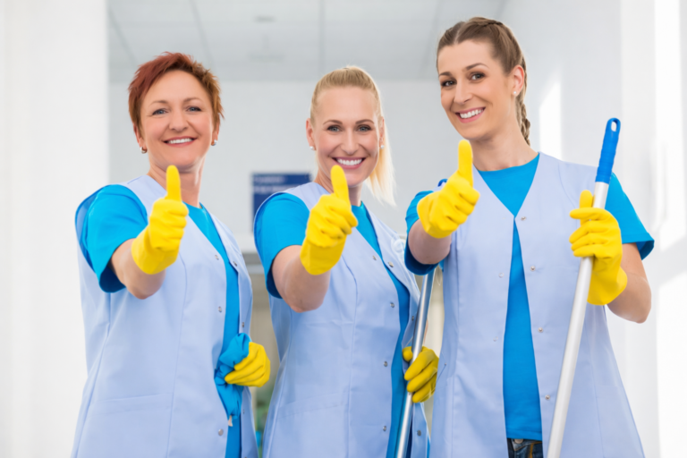 Tre kvinnor i blå uniformer och gula handskar ler och ger tummen upp. De står i en ljus hall och utstrålar positivitet och lagarbete